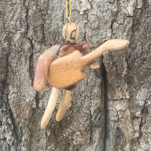 Puede incluir: Un adorno de madera de una persona tocando una guitarra. El adorno está hecho de diferentes tipos de madera, incluyendo una madera marrón clara, una madera marrón oscura y una madera rojiza. El adorno está colgado de una cuerda amarilla.