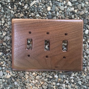 May include: A triple light switch cover made of dark brown walnut wood. The cover has three rectangular cutouts for light switches and three small round holes for screws.