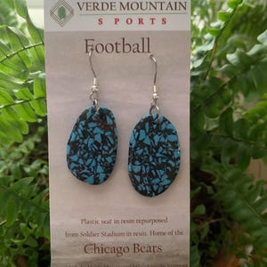 May include: A pair of blue and black football-shaped earrings made from repurposed plastic seats from Soldier Field, home of the Chicago Bears. The earrings are on a white card with the text "Verde Mountain Sports" and "Football".