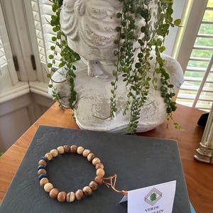 May include: A wooden bead bracelet with natural and blue beads, displayed on a dark grey surface. The bracelet is next to a white planter shaped like a bust, filled with trailing green plants. A tag from Verde Mountain Sports is attached.