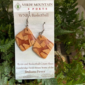 Könnte beinhalten: Diamantförmige Ohrringe mit Holzmaserung. Die Ohrringe bestehen aus Harz und zeigen ein Basketball-Parkett-Design. Die Ohrringe sind von Verde Mountain Sports und stehen in Verbindung mit dem WNBA-Team Indiana Fever.