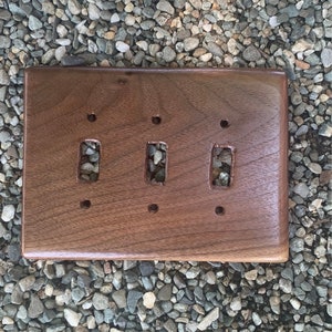 Puede incluir: Una placa de interruptor triple rectangular de madera con un acabado marrón oscuro. Tiene tres recortes rectangulares para los interruptores y cuatro orificios para tornillos. La veta de la madera es visible.