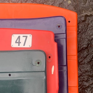May include: A stack of four plastic stadium seats. The top seat is orange, the second is purple, the third is red, and the bottom is dark green. The red seat has a white number 47 on it.