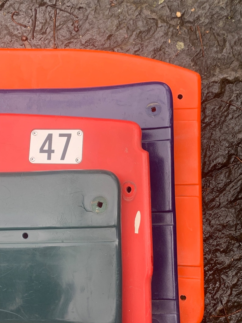 May include: A stack of four plastic stadium seats. The top seat is orange, the second is purple, the third is red, and the bottom is dark green. The red seat has a white number 47 on it.