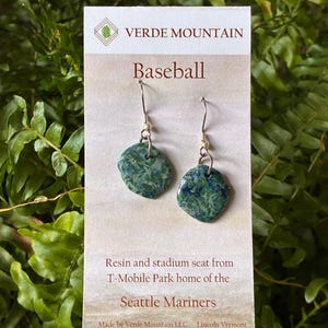 May include: A pair of dangle earrings featuring irregular, mottled green and blue resin charms suspended from silver-toned metal hooks. The earrings are displayed on a white card labeled Verde Mountain, Baseball, and Resin and stadium seat from T-Mobile Park home of the Seattle Mariners.
