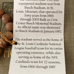 May include: A close-up of a brown wooden bracelet with a white tag. The tag has text that reads: "This bracelet is handmade from the Missouri State Tree and repurposed stadium seat from Busch Stadium. in St. Louis, Missouri, that operated for forty years from 1966 through 2005 Built as Civic Center Busch Memorial Stadium, its official name was shortened to Busch Stadium in January 1982. The stadium served as the home of the St. Louis Cardinals National League baseball team for its entire operating existence, while also serving as home of the NFL Cardinals team for 22 seasons, from 1966 through 1987 www.verdemtn.com"