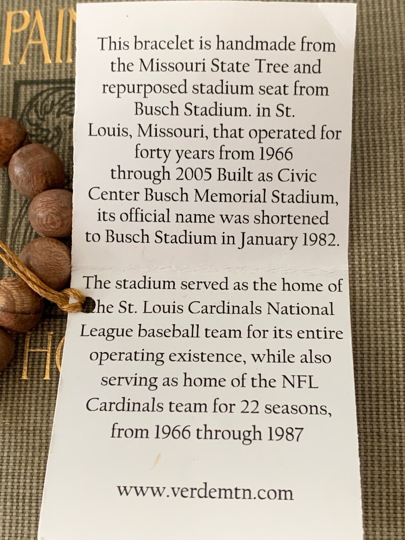 May include: A close-up of a brown wooden bracelet with a white tag. The tag has text that reads: "This bracelet is handmade from the Missouri State Tree and repurposed stadium seat from Busch Stadium. in St. Louis, Missouri, that operated for forty years from 1966 through 2005 Built as Civic Center Busch Memorial Stadium, its official name was shortened to Busch Stadium in January 1982. The stadium served as the home of the St. Louis Cardinals National League baseball team for its entire operating existence, while also serving as home of the NFL Cardinals team for 22 seasons, from 1966 through 1987 www.verdemtn.com"