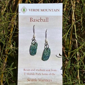 May include: A pair of dangle earrings featuring small, irregular, teal and green speckled resin charms suspended from silver-toned metal hooks. The earrings are displayed on a white card labelled Verde Mountain Baseball, Resin and stadium seat from T-Mobile Park home of the Seattle Mariners.