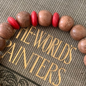 May include: A wooden bracelet with red beads. The bracelet is on a book with the words "The World's Painters" on the cover.