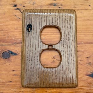 May include: A wooden electrical outlet cover with two openings. The cover is made of light brown wood with a visible grain pattern. The cover is rectangular with rounded edges and is mounted on a wooden surface.