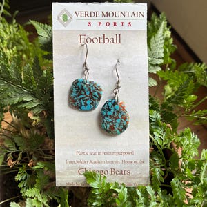 May include: Turquoise and brown resin earrings with silver hooks. The earrings are made from repurposed plastic stadium seats and are labeled "Football" with "Chicago Bears" text. The earrings are displayed on a white card with green accents.