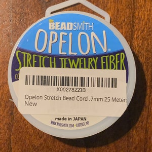 May include: A white spool of Opelon Stretch Jewelry Fiber, a clear elastic bead cord, with a blue and green label that says "the BEADSMITH OPELON STRETCH JEWELRY FIBER" and "X00278ZZIB Opelon Stretch Bead Cord .7mm 25 Meter New made in JAPAN WWW.BEADSMITH.COM CARTERET, NJ".