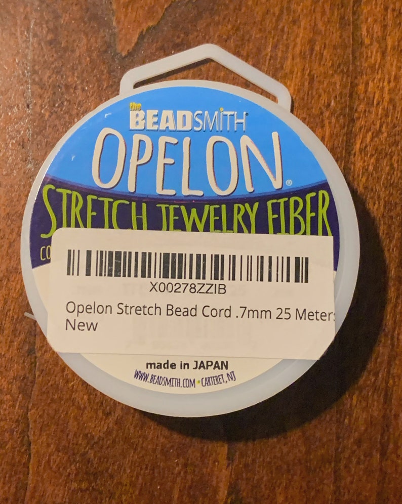 May include: A white spool of Opelon Stretch Jewelry Fiber, a clear elastic bead cord, with a blue and green label that says "the BEADSMITH OPELON STRETCH JEWELRY FIBER" and "X00278ZZIB Opelon Stretch Bead Cord .7mm 25 Meter New made in JAPAN WWW.BEADSMITH.COM CARTERET, NJ".
