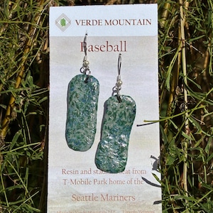 May include: A pair of dangle earrings featuring rectangular, textured resin pendants in shades of green and blue. The earrings are attached to a white display card labeled Verde Mountain, Baseball, and Resin and seat from T-Mobile Park.