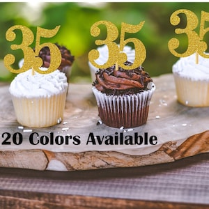Può includere: Tre cupcake con decorazioni a forma di numero 35 in glitter dorati. I cupcake sono glassati con glassa bianca e al cioccolato e hanno codette. I cupcake sono su una superficie di legno. 20 colori disponibili.