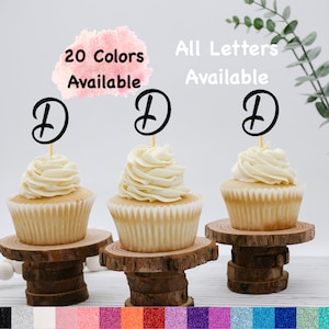 May include: Three cupcakes with white icing topped with black letter D cake toppers. The cupcakes are on wooden cake stands. The text "20 Colors Available" and "All Letters Available" is written above the cupcakes.