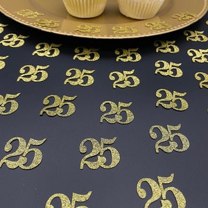 May include: Gold glitter confetti with the number 25 printed on each piece. The confetti is scattered on a black surface. Two cupcakes with white frosting are on a gold plate in the background.