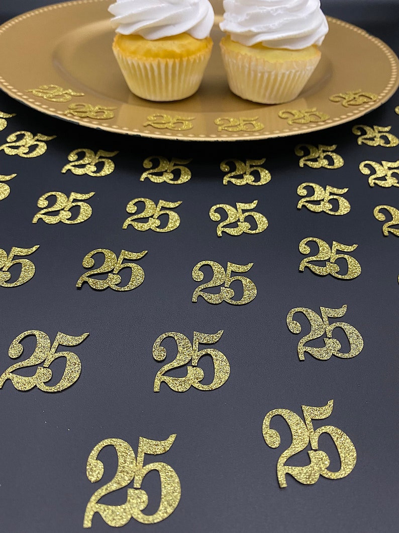 May include: Gold glitter confetti with the number 25 printed on each piece. The confetti is scattered on a black surface. Two cupcakes with white frosting are on a gold plate in the background.