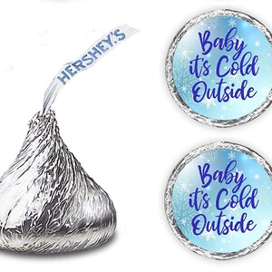 May include: Two blue and silver round stickers with the text "Baby it's Cold Outside" in blue cursive font. The stickers are decorated with white snowflakes. The stickers are designed to be placed on Hershey's Kisses chocolate candies.