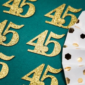 Può includere: Sagome di numeri 45 in glitter dorati su uno sfondo verde. Il numero 45 è ripetuto più volte. Un ventaglio a pois bianchi e dorati si trova nell'angolo in basso a destra dell'immagine.