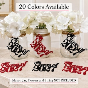 May include: Three clear glass mason jars filled with white artificial flowers, each wrapped with twine and a black or red glittery cutout reading Class of 2021. Three additional Class of 2021 cutouts in red and black glitter lie on the surface.