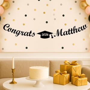 May include: A black banner featuring the text "Congrats" and "Matthew" in a cursive font, separated by a black graduation cap icon with the year "2026" printed in white. The banner is suspended against a wall decorated with gold and black dots.