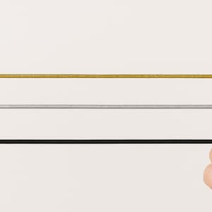 May include: Three thin, flexible elastic cords arranged horizontally. The top cord is metallic gold, the middle cord is metallic silver, and the bottom cord is solid black. Each cord has a smooth, uniform texture and a consistent, narrow diameter.