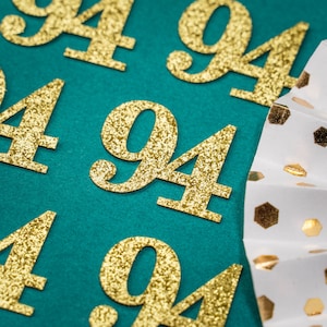 Può includere: Numeri 94 di glitter dorati su uno sfondo turchese. I numeri sono sparsi sulla superficie.