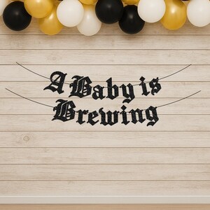 May include: A black glittery banner with the text A Baby is Brewing in a gothic font, suspended by thin black strings. Above the banner, a row of balloons in alternating colors of metallic gold, matte black, and white are arranged in a line.