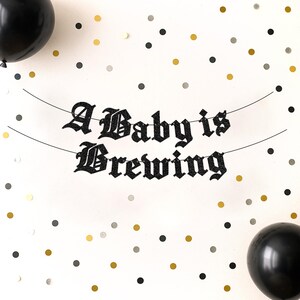 May include: A black glittery banner with the text A Baby is Brewing in a gothic font, suspended on two thin strings. The banner is surrounded by black balloons and scattered gold, black, and silver confetti dots on a plain white background.