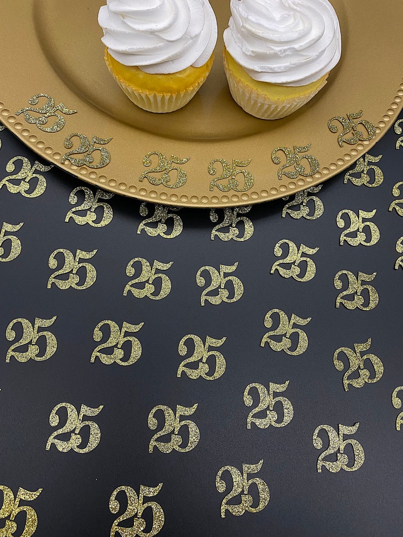 May include: Two cupcakes with white frosting on top of a gold charger. The charger is on a black surface with gold glitter confetti that says "25" scattered around it.