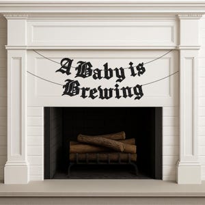 May include: A black glittery banner featuring the text A Baby is Brewing in a gothic font, suspended by thin black strings. The banner is displayed across a white fireplace mantel with a dark interior containing stacked wooden logs.