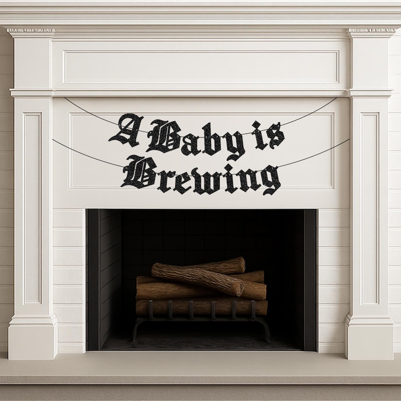 May include: A black glittery banner featuring the text A Baby is Brewing in a gothic font, suspended by thin black strings. The banner is displayed across a white fireplace mantel with a dark interior containing stacked wooden logs.