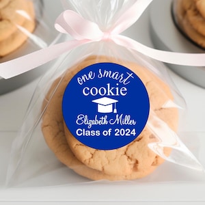 May include: A blue and white round sticker with the text "one smart cookie" and a graduation cap graphic. The sticker is on a clear plastic bag with a pink ribbon and a cookie inside.