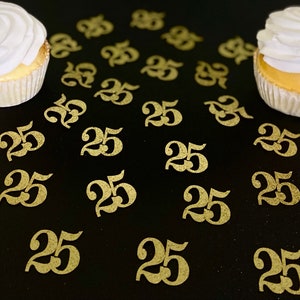 May include: Gold glitter confetti with the number 25 printed on each piece. The confetti is scattered on a black surface with two cupcakes in the background.