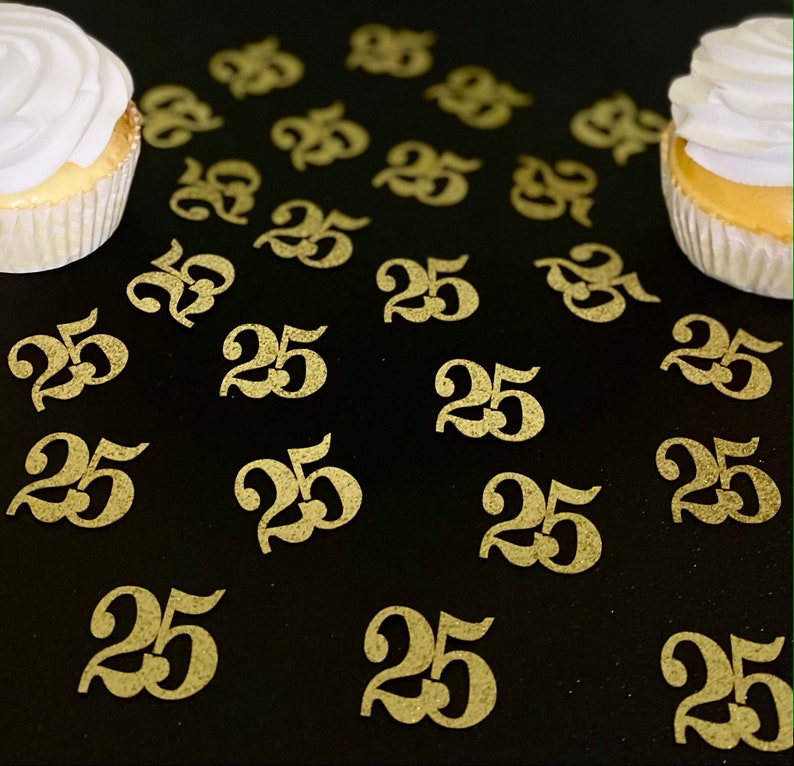 May include: Gold glitter confetti with the number 25 printed on each piece. The confetti is scattered on a black surface with two cupcakes in the background.