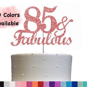 May include: A cake topper with the numbers 85 and the word "fabulous" in sparkly pink glitter. The topper is on a white cake. 20 Colors Available.