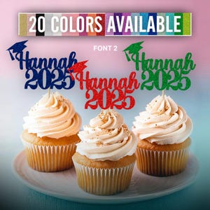 May include: Three cupcakes with graduation themed toppers. The toppers are glittery and say "Hannah 2023" in different colors. The cupcakes are frosted with white frosting and have a light brown cupcake liner.