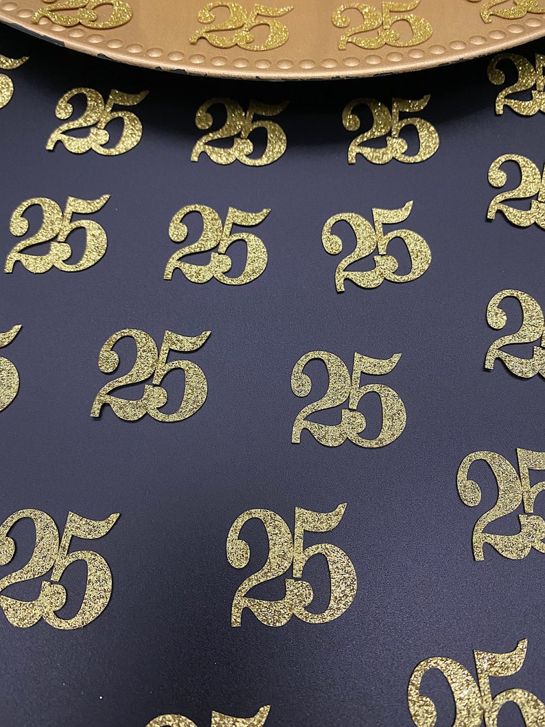 May include: A close-up of a dark blue surface covered with gold glitter-covered number 25s. The numbers are scattered randomly across the surface.