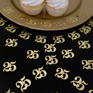May include: Gold glitter number 25 confetti scattered on a black surface with two cupcakes on a gold plate.