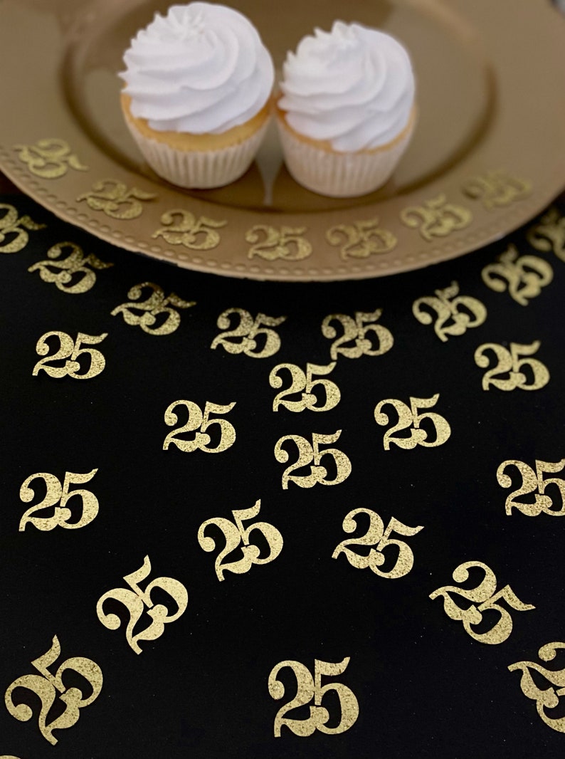 May include: Gold glitter number 25 confetti scattered on a black surface with two cupcakes on a gold plate.