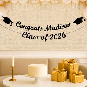 May include: A black banner with the text Congrats Madison Class of 2026 and two graduation cap icons. The banner is displayed above a white cake on a stand, a gold candle holder with a white candle, and three gold gift boxes with ribbons.