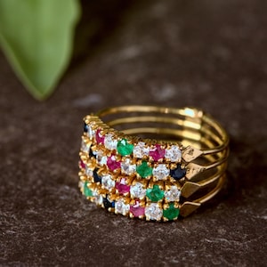 May include: A gold multi-band ring featuring four stacked bands connected at the sides. Each band is set with a row of alternating round-cut gemstones, including clear white stones, vibrant red rubies, deep blue sapphires, and bright green emeralds.