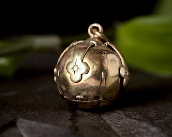 May include: A gold-colored, spherical pendant with a hinged design. The pendant features a four-petal floral design with a central gemstone. The pendant is suspended from a small loop, and is set against a dark background.