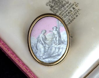 May include: A gold-toned oval brooch with a pink and white cameo featuring two figures. The figures are a woman in a long dress and a young boy with wings. The cameo is set in a gold-toned frame with a beaded edge.