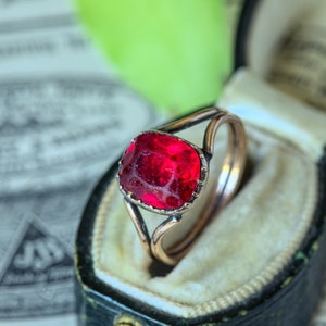 May include: A red gemstone ring set in a gold-toned band. The ring is in a black velvet ring box.