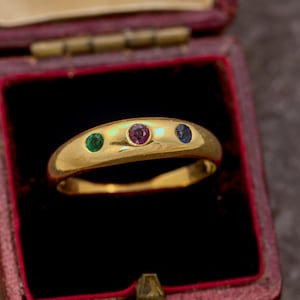 Op de afbeelding: Een gouden ring met drie kleine, ronde, vlak gezette edelstenen die horizontaal zijn gerangschikt. De stenen bestaan uit een groene smaragd aan de linkerkant, een dieproze robijn in het midden en een blauwe saffier aan de rechterkant in gepolijst goud.