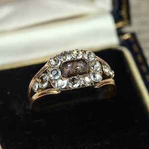 May include: A gold ring with a rectangular center stone surrounded by smaller round stones. The center stone is a dark gray color and the surrounding stones are clear.