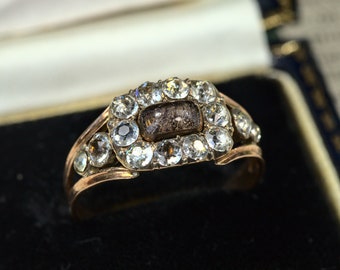 May include: A gold ring with a rectangular center stone surrounded by smaller round stones. The center stone is a dark gray color and the surrounding stones are clear.