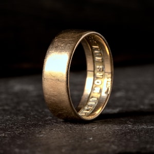 Peut inclure: Une bague dorée avec un extérieur lisse et arrondi. L'intérieur de la bague comporte du texte gravé. La bague est posée sur un fond sombre et texturé, soulignant son éclat métallique. Le design de la bague suggère un style classique et intemporel.